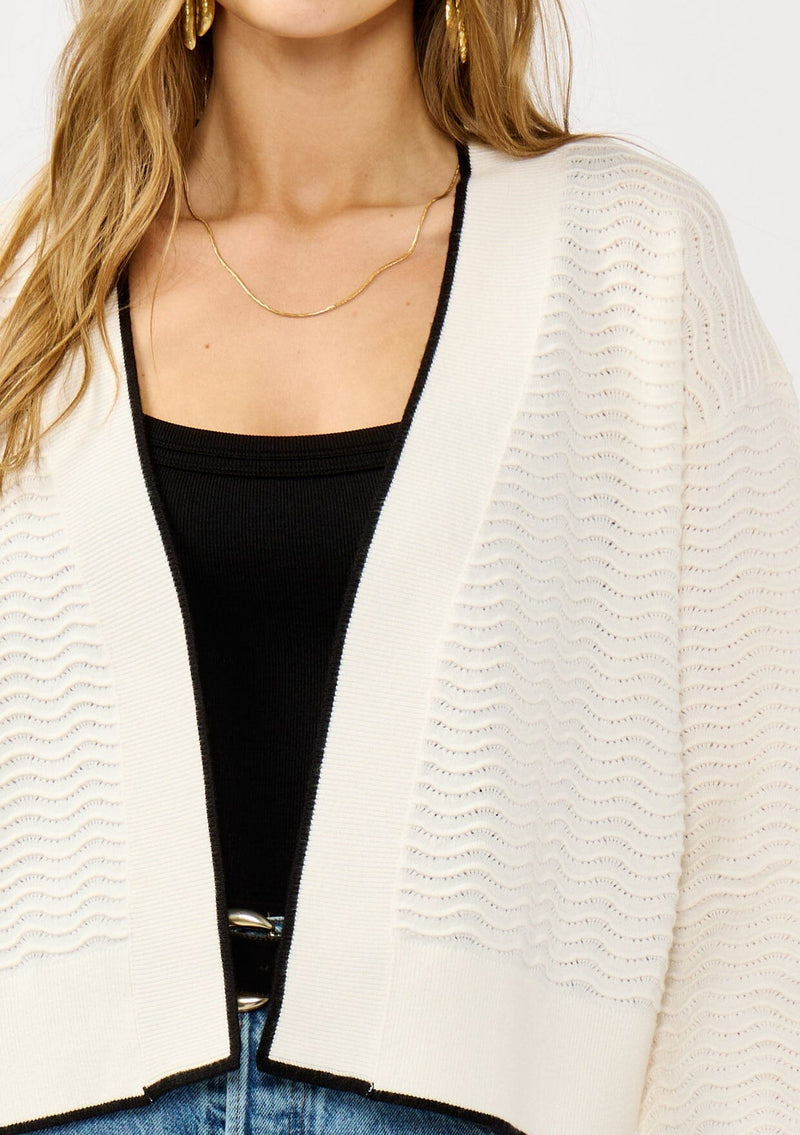 [Color: Vanilla/Black]  A detail image of a blonde model wearing a white pointelle knit cardigan with a black trim. Featuring a textured squiggle pattern, a collarless design, and long sleeves. 