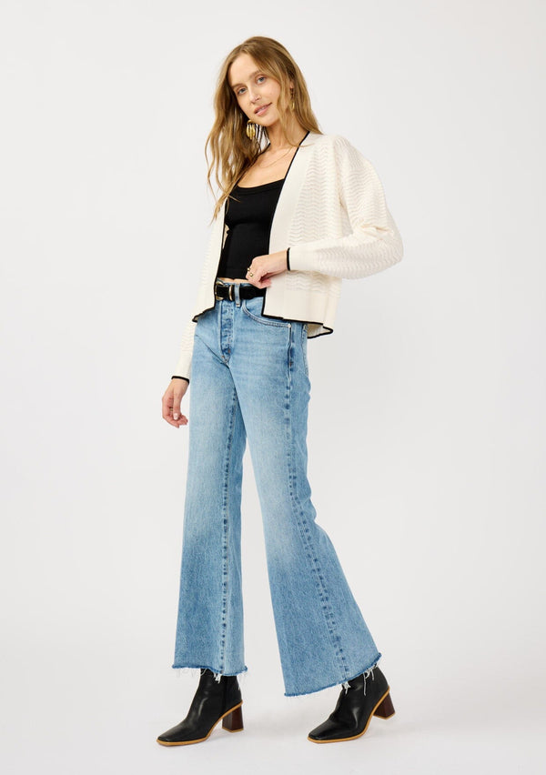 [Color: Vanilla/Black]  A side facing image of a blonde model wearing a white pointelle knit cardigan with a black trim. Featuring a textured squiggle pattern, a collarless design, and long sleeves. 