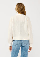 [Color: Vanilla/Black]  A back facing image of a blonde model wearing a white pointelle knit cardigan with a black trim. Featuring a textured squiggle pattern, a collarless design, and long sleeves. 