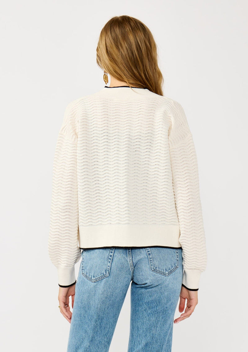 [Color: Vanilla/Black]  A back facing image of a blonde model wearing a white pointelle knit cardigan with a black trim. Featuring a textured squiggle pattern, a collarless design, and long sleeves. 