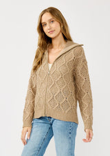 [Color: Mocha] A side facing image of a blonde model wearing a brown cable knit sweater with a full-zip front, funnel neck, and ribbed trims. A classic fall layer to pair with denim. 