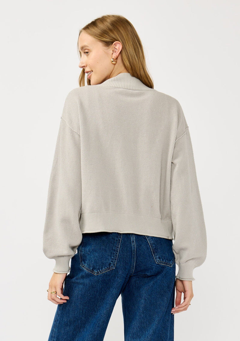 [Color: Heather Light Grey] A back facing image of a blonde model wearing a grey casual, lightweight cardigan with an open front, ribbed trim, and rolled edges. A classic staple cardigan for transitional styling. 