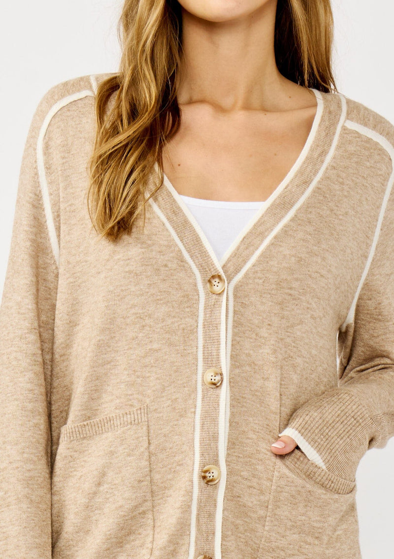 [Color: Taupe/White] A detail image of a blonde model wearing a light brown button front cardigan with white border stripes. Featuring essential patch pockets and a relaxed mid length silhouette. 