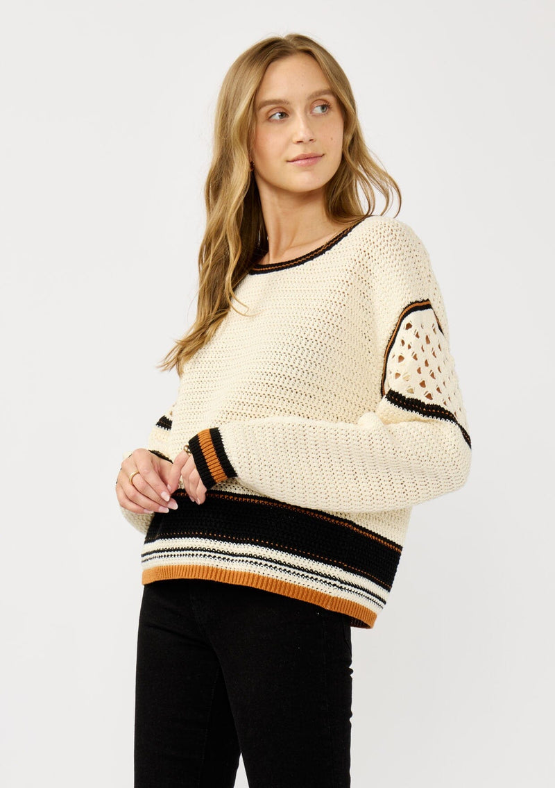 [Color: Natural/Black] A side facing image of a blonde model wearing a cotton pointelle knit sweater with a border stripe. Featuring a classic crew neckline, dropped shoulders, and open knit details along the sleeves. 