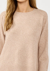 [Color: Heather Oat] A detail image of a blonde model wearing a light brown pullover crew neck sweater with ribbed accents, side vents, and a high low hemline. 