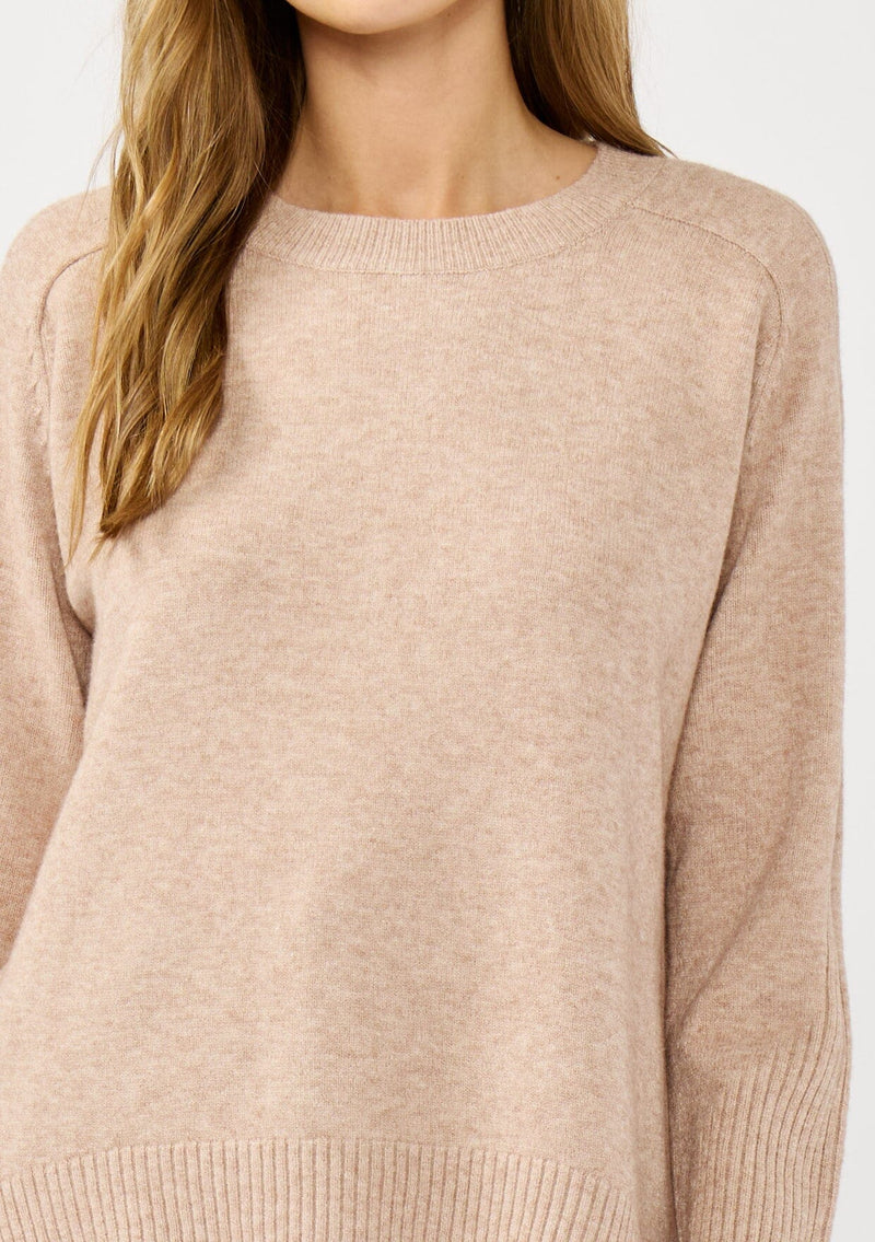 [Color: Heather Oat] A detail image of a blonde model wearing a light brown pullover crew neck sweater with ribbed accents, side vents, and a high low hemline. 