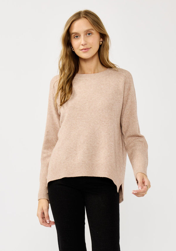 [Color: Heather Oat] A front facing image of a blonde model wearing a light brown pullover crew neck sweater with ribbed accents, side vents, and a high low hemline. 