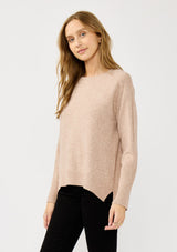[Color: Heather Oat] A side facing image of a blonde model wearing a light brown pullover crew neck sweater with ribbed accents, side vents, and a high low hemline. 