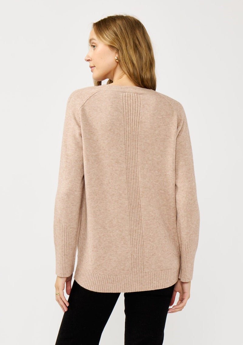 [Color: Heather Oat] A back facing image of a blonde model wearing a light brown pullover crew neck sweater with ribbed accents, side vents, and a high low hemline. 