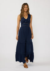 [Color: Navy] A front facing image of a brunette model wearing a flowy navy maxi dress with a halter neck, v-neckline, ruffle details, and a tiered skirt. 