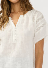 [Color: Off White] A detail image of a brunette model wearing a white cotton gauze top top with lace details on the sleeves and a half button front. 