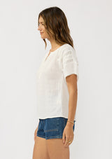 [Color: Off White] A side facing image of a brunette model wearing a white cotton gauze top top with lace details on the sleeves and a half button front. 