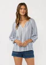 [Color: Blue/ Gold] A front facing image of a brunette model wearing a blue and white striped blouse with gold thread details, a split v-neckline and long sleeves with ruffled cuffs. 