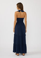 [Color: Navy] A back facing image of a brunette model wearing a flowy navy maxi dress with a halter neck, v-neckline, ruffle details, and a tiered skirt. 