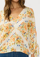 [Color: Ivory/Coral] A detail image of a brunette model wearing an orange floral blouse with voluminous long sleeves, a tie detail, and v-neckline. 