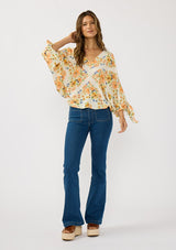 [Color: Ivory/Coral] A front facing image of a brunette model wearing an orange floral blouse with voluminous long sleeves, a tie detail, and v-neckline. 