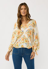 [Color: Ivory/Coral] A front facing image of a brunette model wearing an orange floral blouse with voluminous long sleeves, a tie detail, and v-neckline. 