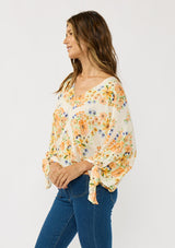 [Color: Ivory/Coral] A side facing image of a brunette model wearing an orange floral blouse with voluminous long sleeves, a tie detail, and v-neckline. 