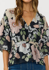 [Color: Black/Blush] A detail image of a brunette model wearing a black and pink floral blouse with a v-neckline, adjustable drawstring waist, and flowy half length sleeves with ruched shoulder detail. 