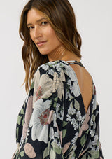 [Color: Black/Blush] A detail image of a brunette model wearing a black and pink floral blouse with a v-neckline, adjustable drawstring waist, and flowy half length sleeves with ruched shoulder detail. 