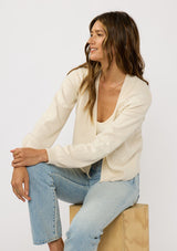 [Color: Ivory] A sitting image of a brunette model wearing an ivory cardigan with a collarless design, floral embroidery, and rib trims. 