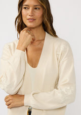 [Color: Ivory] A front facing image of a brunette model wearing an ivory cardigan with a collarless design, floral embroidery, and rib trims. 
