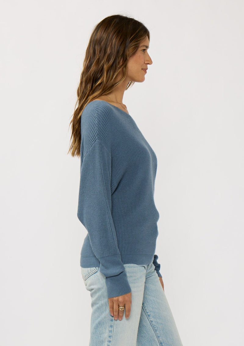 [Color: Heather Ocean] A side facing image of a brunette model wearing a blue waffle knit pullover sweater. With long sleeves, a relaxed fit, and a wide neckline that can be worn off the shoulder.