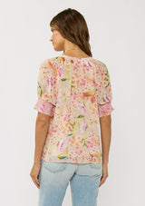 [Color: Natural/Fuchsia] A back facing image of a brunette model wearing a pink floral top with a button front and puff sleeves with smocked cuffs. A pretty spring casual top paired with denim. 