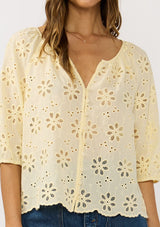 [Color: Yellow] A detail image of a brunette model wearing a floral eyelet embroidered top with half sleeves, a button front, and scallop trimmed hem. 