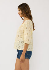 [Color: Yellow] A side facing image of a brunette model wearing a floral eyelet embroidered top with half sleeves, a button front, and scallop trimmed hem. 