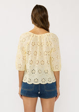 [Color: Yellow] A back facing image of a brunette model wearing a floral eyelet embroidered top with half sleeves, a button front, and scallop trimmed hem. 