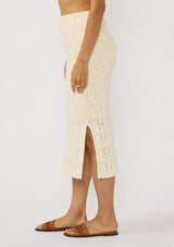 [Color: Natural] A side facing image of a brunette model wearing a beige pointelle knit midi skirt with side slits and a ribbed elastic waistband. 