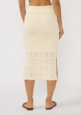 [Color: Natural] A back facing image of a brunette model wearing a beige pointelle knit midi skirt with side slits and a ribbed elastic waistband. 