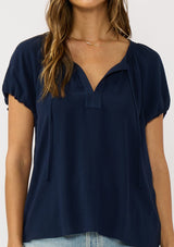 [Color: Navy] A detail image of a brunette model wearing a navy blue puff sleeve top with a split v-neckline and neck ties. 