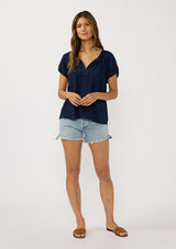 [Color: Navy] A front facing image of a brunette model wearing a navy blue puff sleeve top with a split v-neckline and neck ties. 