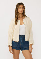 [Color: Natural] A front facing image of a brunette model wearing a beige jacquard jacket with a collarless design, zip-up front, and side pockets. A lightweight jacket for the spring season. 