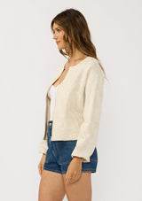 [Color: Natural] A side facing image of a brunette model wearing a beige jacquard jacket with a collarless design, zip-up front, and side pockets. A lightweight jacket for the spring season. 