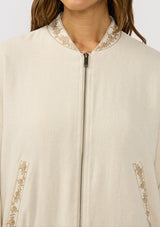 [Color: Natural] A detail image of a brunette model wearing a beige embroidered jacket with a dropped shoulder, zipper front, and a side pockets. 