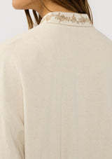 [Color: Natural] A detail image of a brunette model wearing a beige embroidered jacket with a dropped shoulder, zipper front, and a side pockets. 
