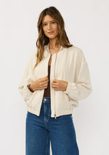[Color: Natural] A front facing image of a brunette model wearing a beige embroidered jacket with a dropped shoulder, zipper front, and a side pockets. 