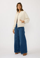 [Color: Natural] A side facing image of a brunette model wearing a beige embroidered jacket with a dropped shoulder, zipper front, and a side pockets. 