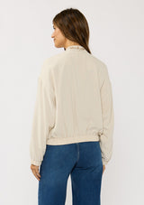[Color: Natural] A back facing image of a brunette model wearing a beige embroidered jacket with a dropped shoulder, zipper front, and a side pockets. 