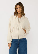 [Color: Natural] A front facing image of a brunette model wearing a beige embroidered jacket with a dropped shoulder, zipper front, and a side pockets. 