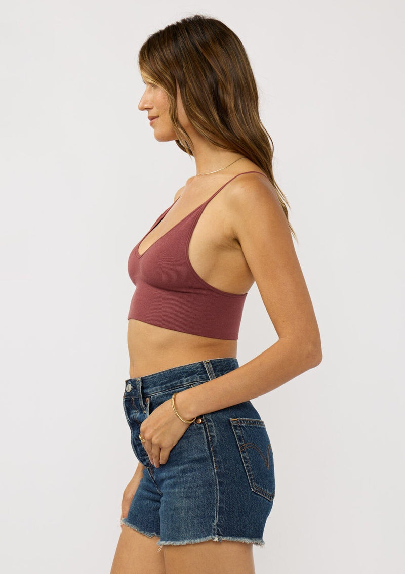 [Color: Burgundy] A  side facing image of a brunette model wearing a basic burgundy bralette cami with a slim stretchy fit and a v-neckline.