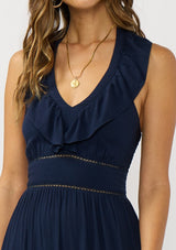 [Color: Navy] A detail image of a brunette model wearing a flowy navy maxi dress with a halter neck, v-neckline, ruffle details, and a tiered skirt. 