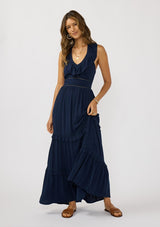 [Color: Navy] A front facing image of a brunette model wearing a flowy navy maxi dress with a halter neck, v-neckline, ruffle details, and a tiered skirt. 
