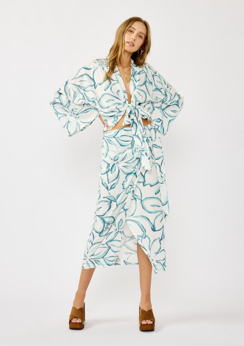 [Color: Ivory/Dark Teal] A front facing image of a blonde model wearing a white and teal tropical floral print wrap skirt, tie waist, and a smocked back. Styled with a matching tie front top. Perfect for vacation or casual outings. 