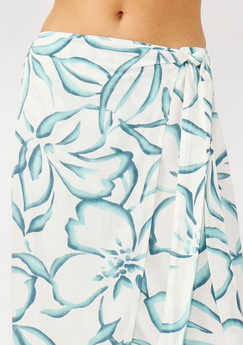 [Color: Ivory/Dark Teal] A detail image of a blonde model wearing a white and teal tropical floral print wrap skirt, tie waist, and a smocked back. Styled with a matching tie front top. Perfect for vacation or casual outings. 