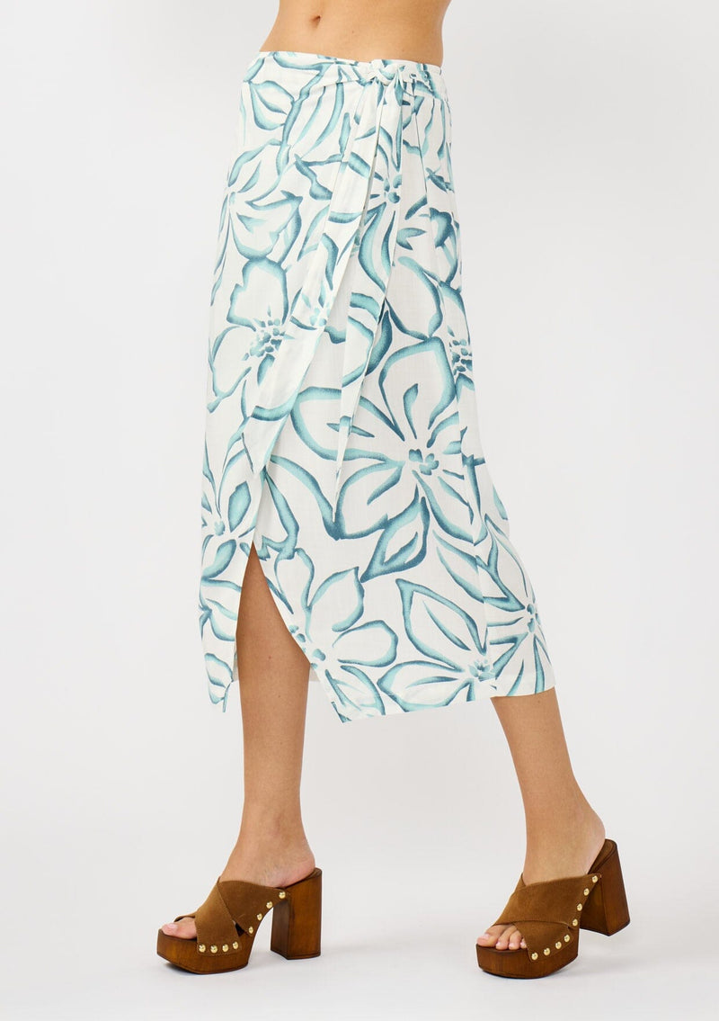 [Color: Ivory/Dark Teal] A side facing image of a blonde model wearing a white and teal tropical floral print wrap skirt, tie waist, and a smocked back. Styled with a matching tie front top. Perfect for vacation or casual outings. 
