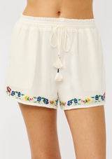 [Color: Cream] A front facing image of a cream floral embroidered short crafted in soft cotton. Featuring a smocked waist, a drawstring, and side pockets. 
